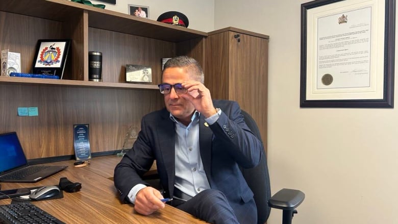 Man in a blue suit and dark glasses sits at a wooden desk. 