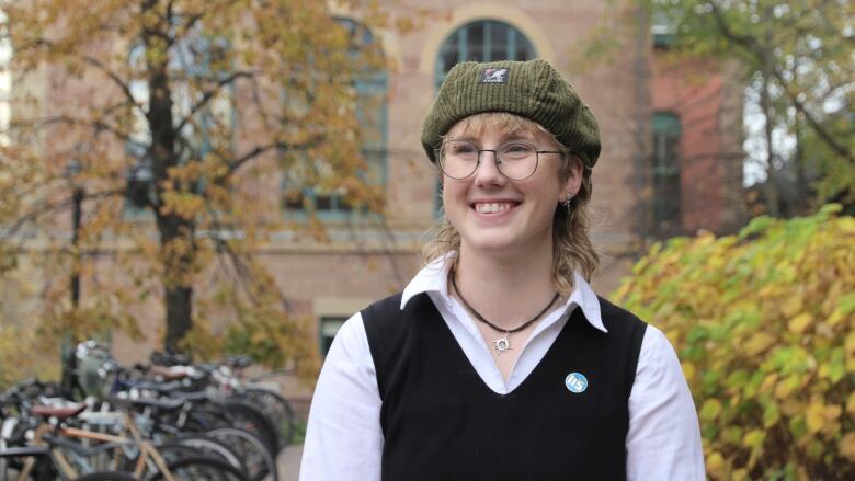 A smiling person wearing glasses, a collared shirt, a sweater-vest and a hat stand in front of a brick building and bicycles. It is fall. 