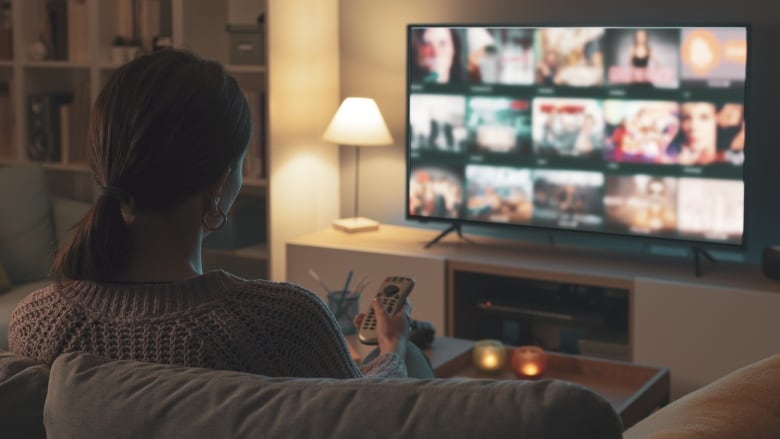 Woman relaxing on the couch, she is using the remote control and choosing a TV show or movie on the television menu