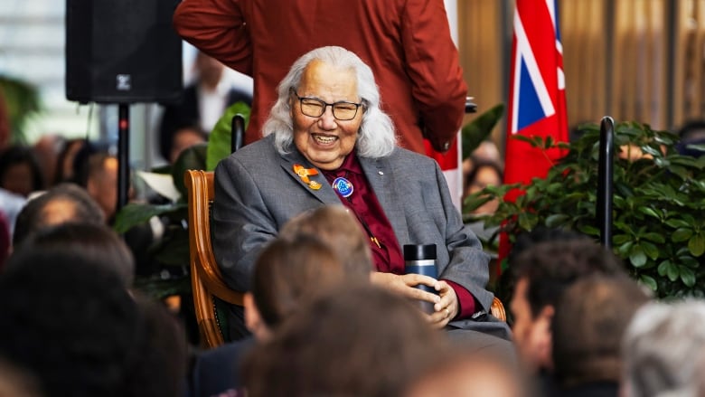 A man on a stage sitting in a chair smiles, with a crowd in front of him.