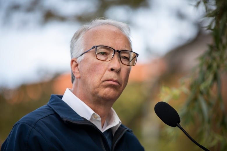 A white man looks to his right while speaking to a mic outdoors.