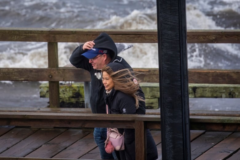 Two people struggle to hold onto their hats and hoodies as they walk on a pier.