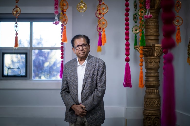 An Indian man stands in front of decorations with his hands clasped beneath his torso.