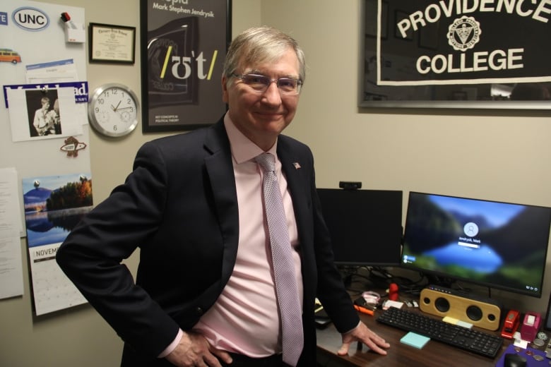 A man in a black blazer with a pink shirt underneath stands in his office, with his left hand placed on his desk.