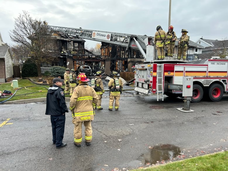 Firefighters stand in the street in front of a burned out house