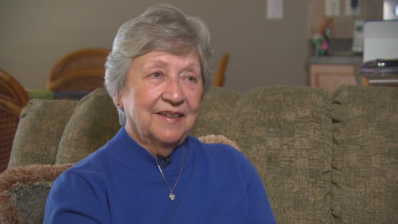 An older woman, with short, wavy grey hair, wears a blue sweater and looks off to the side of the camera.