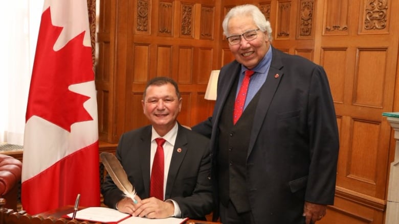 A seated man holds an eagle feather while another stands with his own on his should.