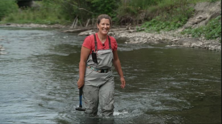 A woman in gray overall waders and a red t-shirt wades through knee deep water in the middle of a river carrying something that looks like a mallet. She's smiling and has her hair pulled back. The riverbank behind her has rocks and greenery.