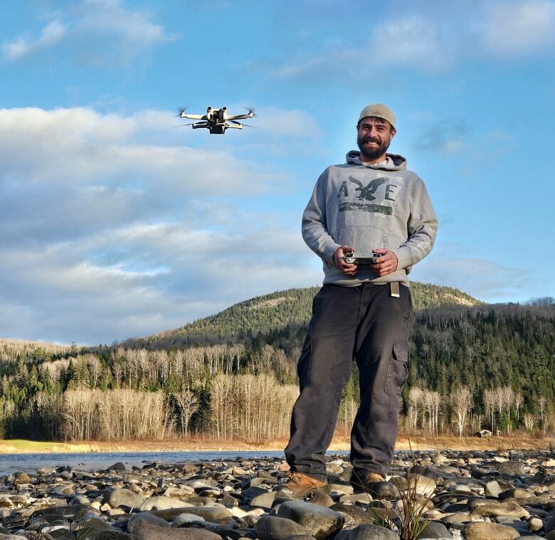 A man in his late 30s or early 40s wearing a gray sweatshirt, ball cap, black pants and work boots stands on a rocky river shoreline operating a drone just above his head. A river, bare deciduous trees, evergreens, hills and and a blue sky with a few gray clouds can be seen in the background.