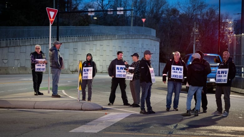 A line of men stand on the road and a traffic island at an intersection, all wearing signs that say 'ILWU Local 514 Locked Out.' It is nighttime. 