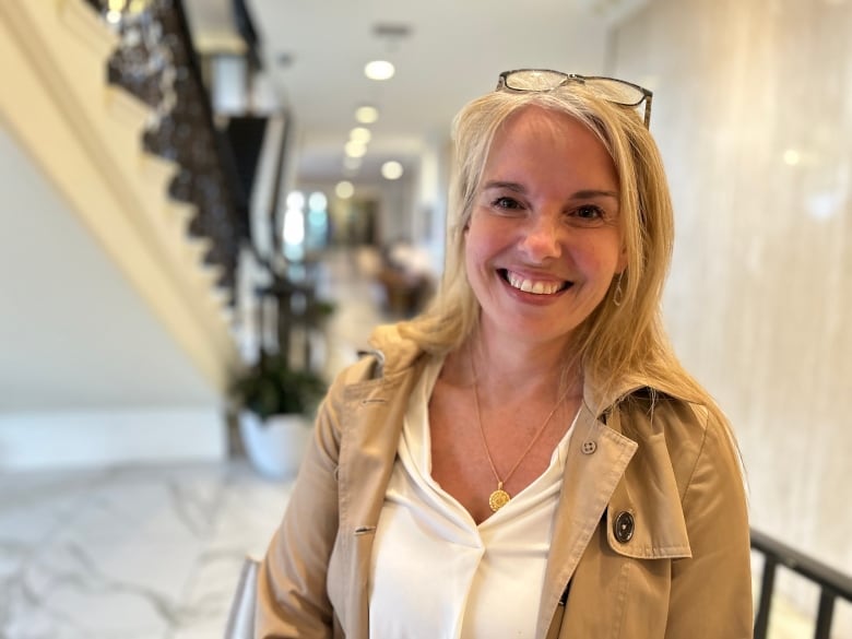 A blonde woman with glasses on her head smiles at the camera, wearing a tan trench coat. 