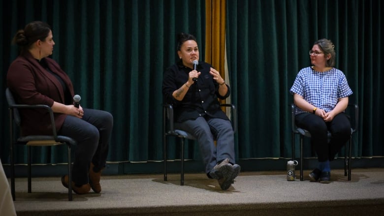 Three women sit on chairs in front of a green curtain, the woman in the middle has a microphone while the others listen.