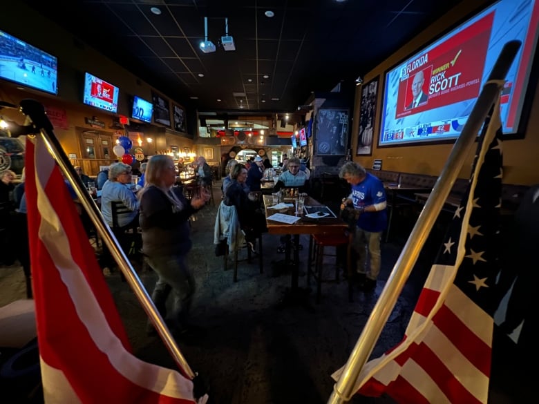 A bar with a few people in it with two American flags in the foreground and a man with a blue Democrats Abroad shirt at a table talking to people