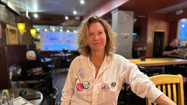 A portrait of a person in a restaurant wearing multiple campaign buttons on their button-up shirt. 