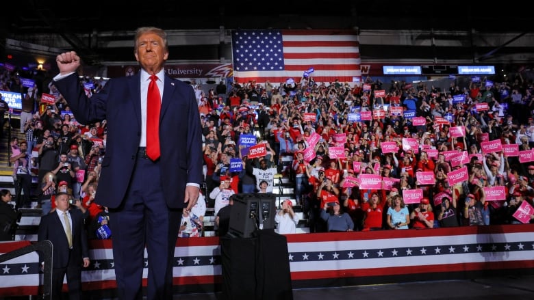 A man in a suit with a red tie holds his fist in the air as dozens of people behind him cheer.