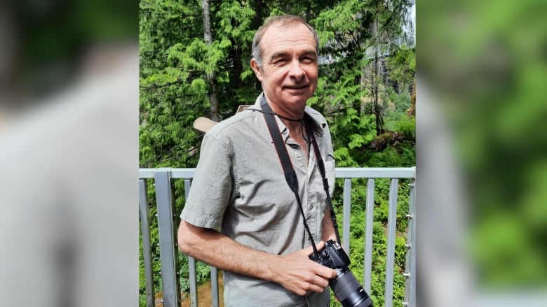 A man holding a camera in front of a forested environment smiles.