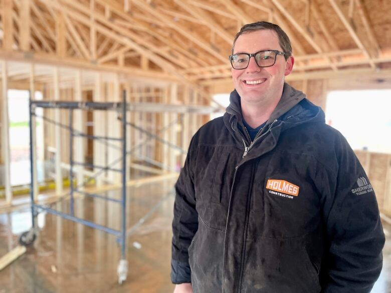 A man with short hair and glasses wearing a dark jacket with a logo reading 'Holmes Construction' standing in a home that's under construction.