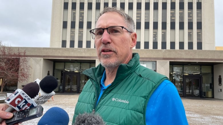 A man stands in front of Regina city hall on Nov. 6, 2024. The man is wearing a blue shirt and a green vest as he speaks into microphones. 