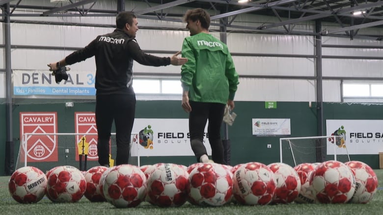 Soccer balls are pictured on a turf field with two people, one wearing black and one wearing green in the background.