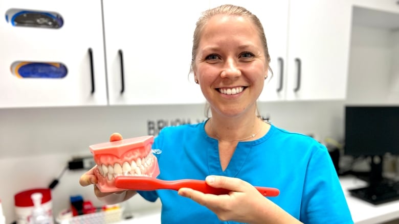 A woman wearing a blue uniform while smiling holds up a giant sized toothbrush and teeth. 