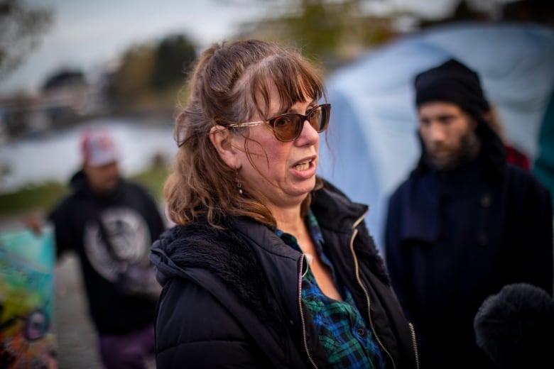 A woman wearing sunglasses speaks in an outdoor park as others look on.