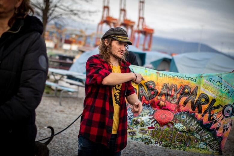 A man wearing a cap and a checked top points to a colourful banner saying 'Unsurrendered ABAY'X PARK'.