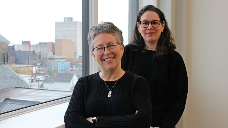 Two women wearing black stand in front of a window smiling.