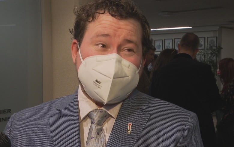 A person in a light blue suit wearing a surgical mask stands in a crowded room.