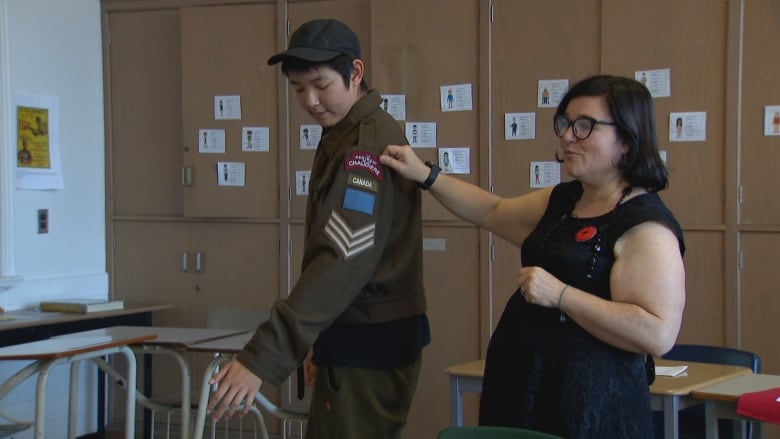 A male student wearing a brown miltary jacket with a regimental badge and chevrons stands in a classroom as a female teacher gestures to the badges on his arm. 