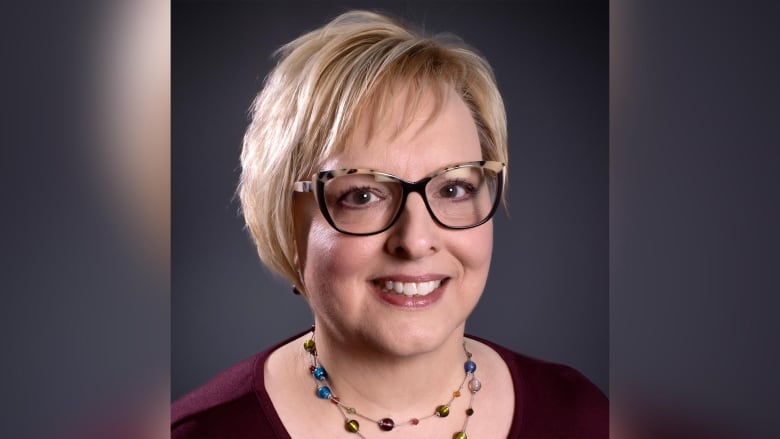 A portrait of a smiling women with short blond hair, tortoiseshell glasses and a necklace of colourful stones.