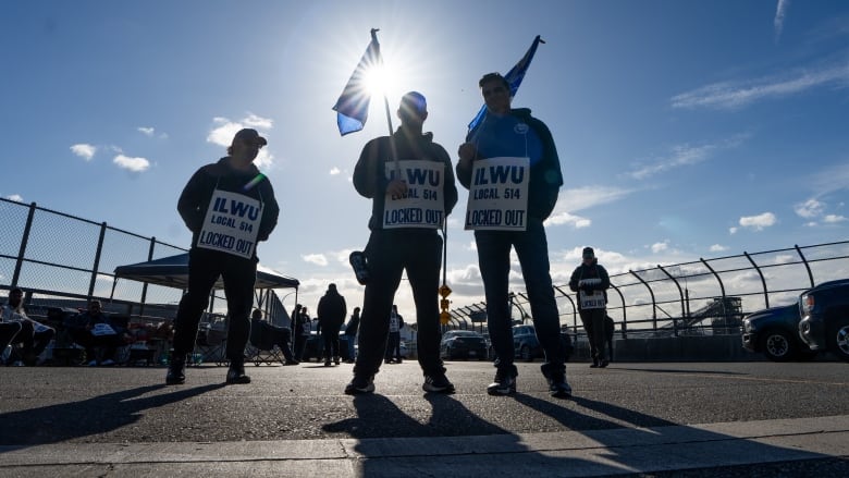 Three people wearing protest signs face the camera against the backdrop of a blue, sunny sky. The signs read 