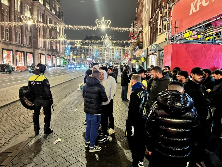 A police officer with a baton walks past a line of young men on an urban street.