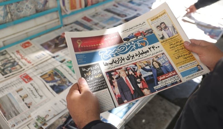 A newspaper is shown, being held up by someone whose hands are also visible. It's being held above a table covered in other newspapers. 