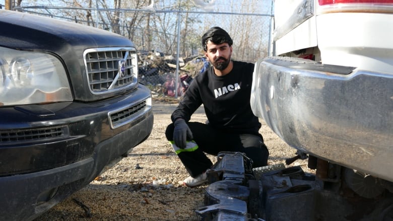 A man is crouching down between the bumpers of two vehicles. He is outside.