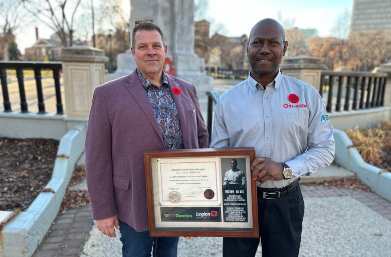 Two men stand next to each other with a framed certificate in front of a cenotaph