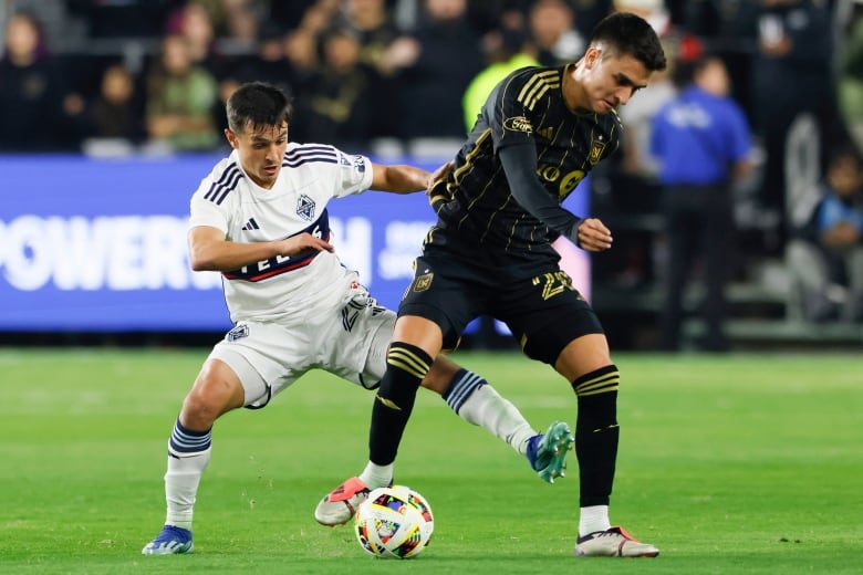 A soccer player holds onto the ball as another player tries to take it from behind.