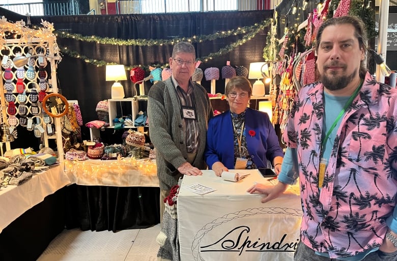 A middle-aged couple and a young man stand near each other inside a booth full of knitting crafts such as winter hats and mittens. They are leaning over a table with their company's logo: Spindrift.