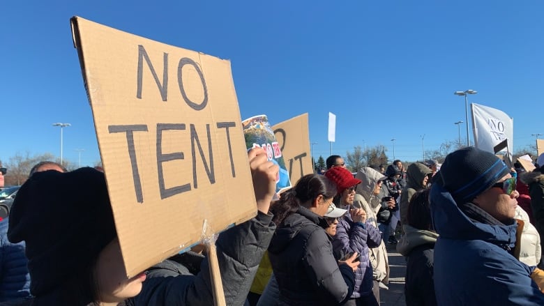 A crowd of people stand together outside. Some of them hold signs, with one cardboard sign in the foreground reading 'No Tent.'