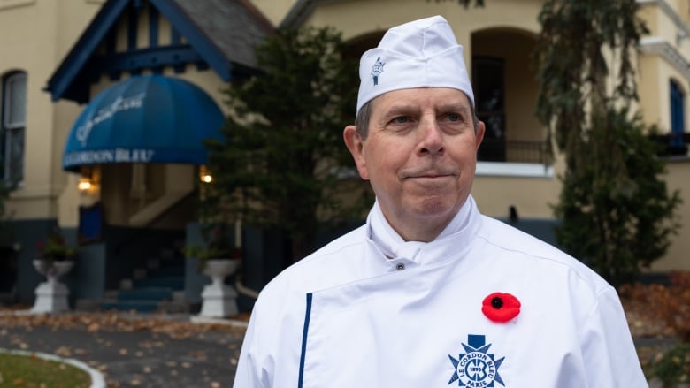 A man stands outside an ornate building on a cloudy day, wearing a chef's uniform and a red poppy.