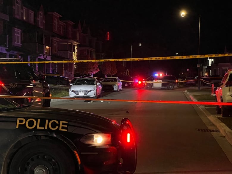 Police cars are seen behind crime scene tape, alongside parked cars on the street. 