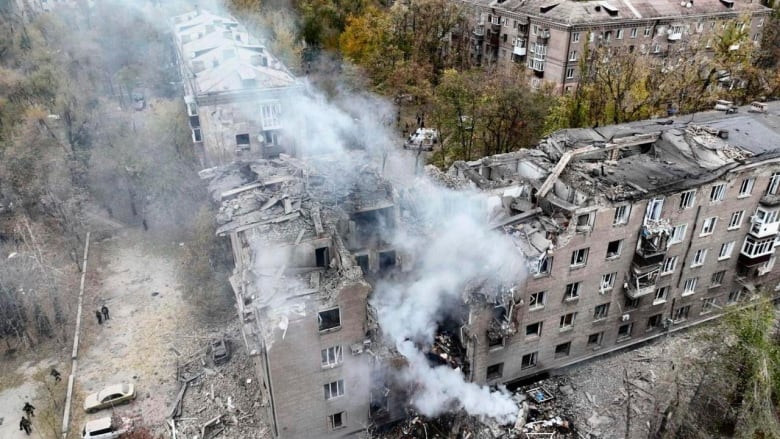 An aerial shot shows the the corner of a street and a five storey apartment complex with a large hole in it, from which smoke is rising. Around the hole, there are crumbling bits of the building and there is debris on the ground near the building as well. 