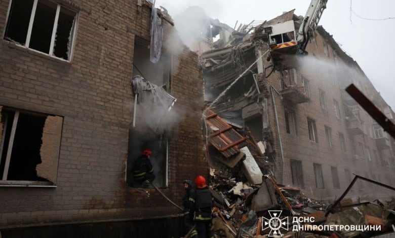 A shot taken by someone standing on the street shows an upward view of a building that has sustained damage. Half of the building is crumbling, with debris piled at the bottom. A crane is visible approaching the top of the affected building. A brick building is on the other side of the image. 