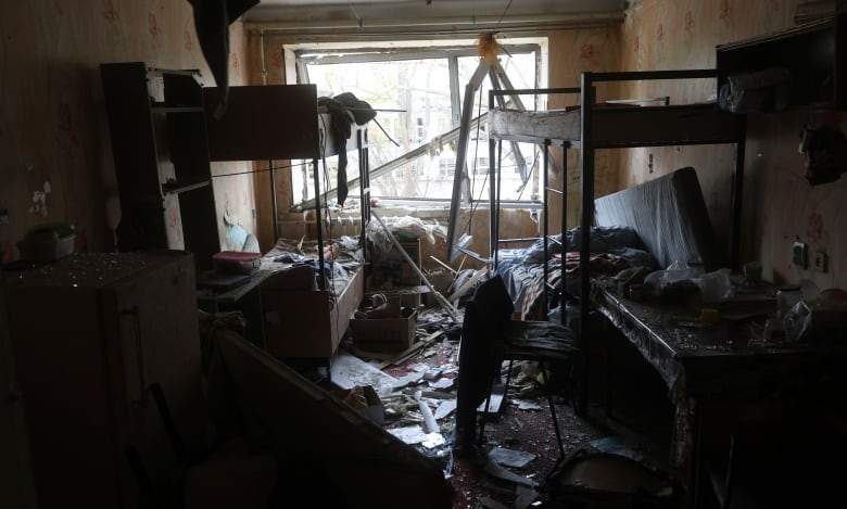 Taken from inside a ruined apartment, the photo shows a darkened bedroom filled with debris and two bunkbeds. There's a smashed window at the far end of the room, through which light is entering. 