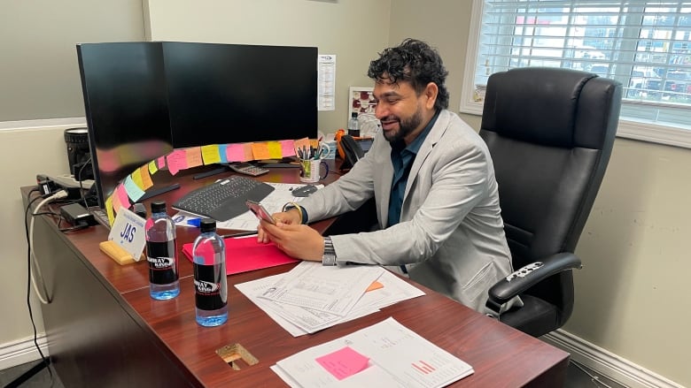 A man with dark hair and a bears sits at a desk with two large computer screens covered in colourful post-it notes while checking his phone. 