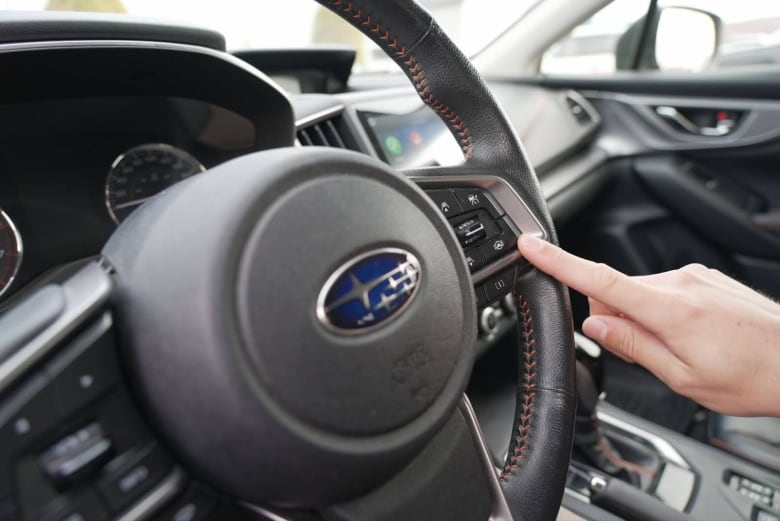 An index finger points to some buttons on a Subaru car steering wheel. 