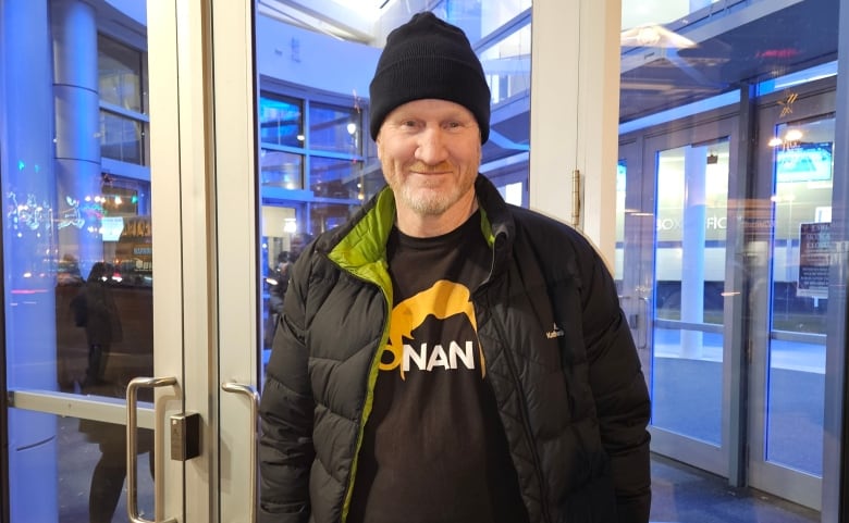 A man in a black jacket and black tuque smiles and stands in front of a door