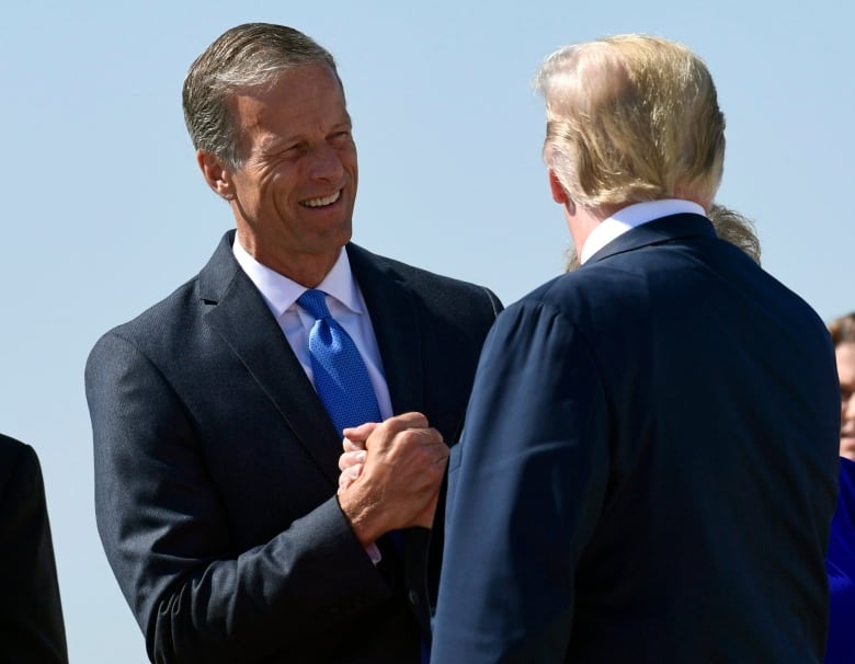 A cleanshaven man in a suit and tie clasps hands with another man in a suit jacket who has his back to the camera.