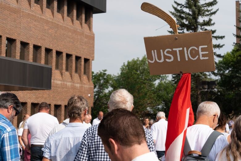 Protesters face away from the camera. One holds a hockey stick covered by a Canadian flag and a cardboard sign that reads, 