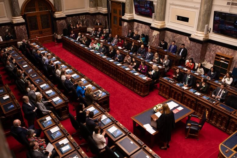 A group of MLAs listen to a clerk delivering instructions.