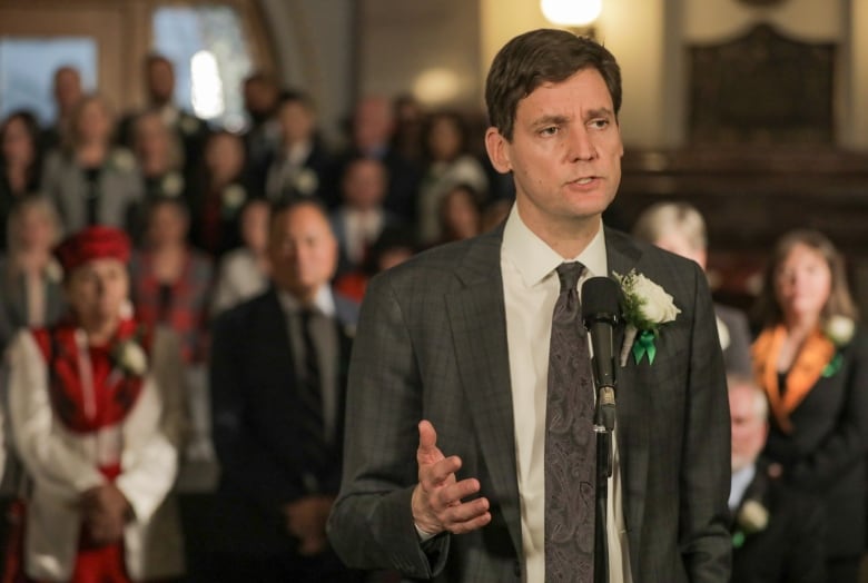 A man speaks into a microphone with a group of people out of focus in the background. 